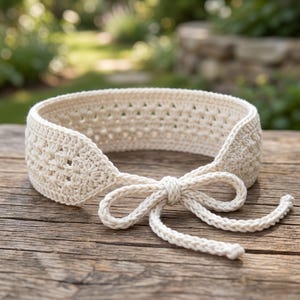 Peut inclure: Bandeau crocheté de couleur crème avec un nœud. Le bandeau présente un motif texturé avec des détails ajourés. Le nœud est centré à l'avant, et le bandeau est posé sur une surface en bois. L'arrière-plan est flou, avec de la verdure visible.