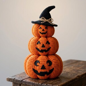 Puede incluir: Una pila de tres calabazas de ganchillo naranjas con caras de jack-o'-lantern de fieltro negro. La calabaza superior lleva un sombrero de bruja de ganchillo negro adornado con un pequeño lazo de cuerda beige. Las calabazas están apiladas verticalmente.