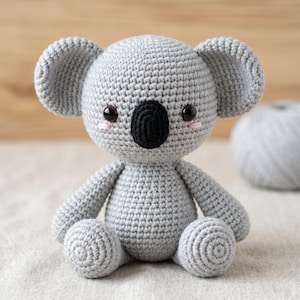 May include: A grey crocheted koala stuffed animal with a large round head, prominent ears, and a black oval nose. The toy features dark brown safety eyes and small pink stitched cheeks, with a textured yarn surface and a seated posture.