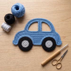 May include: A blue crocheted car applique with black and gray wheels. The set includes three balls of yarn in blue, black, and white, a wooden-handled crochet hook, and small gold-toned scissors with intricate handle detailing for craft projects.