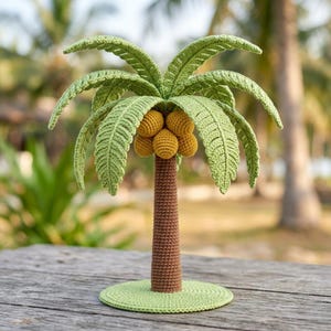 May include: A crocheted palm tree featuring a brown textured trunk, a circular light green base, and several long, pointed green fronds. A cluster of four round, golden-yellow crocheted coconuts is attached to the centre of the tree trunk.