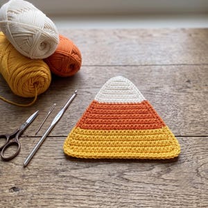May include: A triangular crochet piece featuring horizontal stripes of white, orange, and yellow, resembling a candy corn. Beside it are three skeins of yarn in matching colors, a metal crochet hook, a sewing needle, and a pair of small scissors.