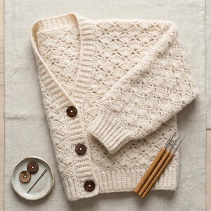 Könnte beinhalten: Cremefarbene gehäkelte Strickjacke mit strukturiertem Muster und drei großen, runden, braunen Knöpfen. Der Pullover hat einen V-Ausschnitt und gerippte Bündchen. Ein kleiner Teller mit zwei Holzknöpfen und einer Nadel sowie drei Häkelnadeln sind ebenfalls sichtbar.
