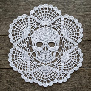 May include: A white circular crochet doily featuring a central skull motif. The design is composed of intricate threadwork with a detailed human skull at the center, surrounded by a radiating pattern of openwork stitches and a scalloped edge border.