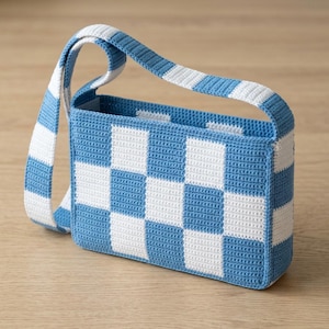 May include: A rectangular shoulder bag crafted with a crochet technique, featuring a repeating blue and white checkered pattern. The bag includes a matching wide shoulder strap with the same blue and white block design, constructed with a dense stitch.