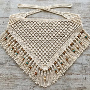 May include: A cream-colored triangular crochet bandana featuring an open diamond-shaped mesh pattern. The bottom edge is finished with long fringe tassels accented by small wooden and turquoise-colored beads, with a small crochet flower at the tip.