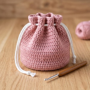 Könnte beinhalten: Ein handgefertigter rosa Häkelbeutel mit Kordelzug und weißer Kordel. Ein Häkelhaken und ein Knäuel rosa Garn liegen auf einer Holzoberfläche. Die Tasche ist rund und hat eine strukturierte Optik.