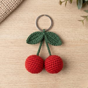 Puede incluir: Un llavero de ganchillo hecho a mano con dos cerezas rojas con hojas y tallos verdes, sujeto a un anillo de metal plateado. Las cerezas son redondas y texturizadas. El llavero está sobre una superficie de madera clara.