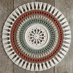 May include: A circular crochet doily featuring a central white starburst pattern, surrounded by concentric rings of sage green, beige bobble stitches, and a rust-coloured band. The outer edge is finished with a fringe of white yarn tassels.