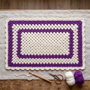 May include: A rectangular crochet mat featuring a granny square pattern with alternating bands of cream and deep purple yarn. The mat is finished with a scalloped cream border. A wooden crochet hook, metal scissors, and two yarn balls are included.