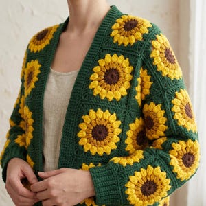 May include: A handmade, open-front cardigan featuring a sunflower design. The crochet sweater has a dark green base with yellow and brown sunflower motifs. The sleeves and body are adorned with the floral pattern, creating a cozy and vibrant garment.