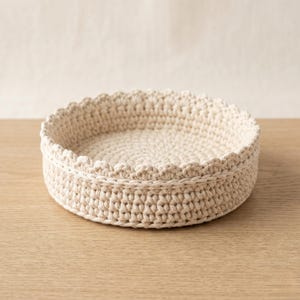 Op de afbeelding: Een ronde, crèmekleurige gehaakte mand met een geschulpte rand. De mand is gemaakt van dik garen en staat op een lichtgekleurd houten oppervlak. Het ontwerp van de mand is eenvoudig en functioneel, geschikt voor opslag of weergave.