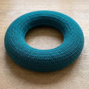 May include: A circular, donut-shaped object covered in a dense, teal-colored crochet stitch pattern. The item features a consistent, textured yarn surface throughout its entire rounded form, creating a uniform and solid appearance in a deep blue-green hue.