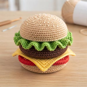 May include: A crocheted hamburger toy featuring a tan bun with white embroidered sesame seeds, a ruffled green lettuce layer, a dark brown patty, a yellow triangular slice of cheese, and a red tomato slice atop a tan bottom bun.