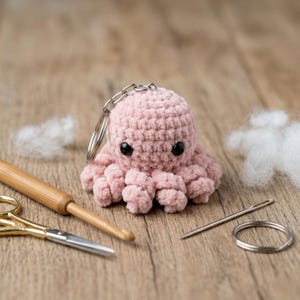 May include: A handmade pink crochet octopus keychain with black safety eyes and a silver keyring. The octopus is surrounded by crafting tools, including scissors, a crochet hook, a needle, and stuffing on a wooden surface.
