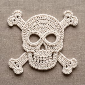 May include: A cream-colored crochet skull and crossbones applique. The design features a central skull with two large eye sockets, a small nasal cavity, and a row of teeth, with two crossed bones extending from behind the skull in a classic motif.