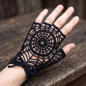 May include: A black fingerless glove crafted from crochet thread, featuring an intricate spiderweb pattern that radiates from a central circular motif. The design includes a delicate tie closure at the wrist, leaving the fingers and thumb exposed.