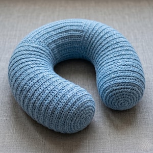 May include: A U-shaped neck pillow crafted from light blue yarn with a visible crochet stitch pattern. The pillow features a consistent ribbed texture throughout its curved design, providing a soft and uniform appearance for neck support.