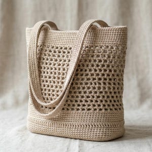 May include: A beige crochet tote bag with two long handles. The bag features a textured, open weave design on the main body and a solid crochet base and top. The bag is made of a soft, natural-looking yarn.