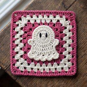 May include: A handmade crochet coaster featuring a white ghost applique on a pink and white granny square. The coaster is square-shaped and rests on a wooden surface. The ghost has two black eyes.