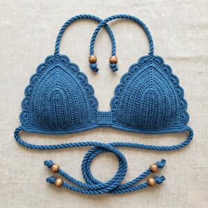May include: A blue crochet bikini top with scalloped edges and rope straps. The top features wooden bead accents and a matching rope tie. The handmade swimwear is displayed on a neutral linen surface.