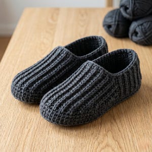 May include: A pair of dark charcoal gray slippers crafted with a thick, ribbed crochet knit pattern. The slippers feature a rounded toe design and a low-cut opening, showcasing a consistent textured yarn construction throughout the entire footwear.