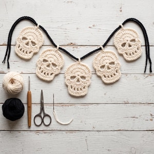 May include: A crochet garland featuring five off-white skull motifs strung on a black yarn cord. Included are a ball of off-white yarn, a ball of black yarn, a wooden crochet hook, a pair of metal scissors, and a metal sewing needle with yarn.