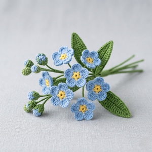May include: A close-up of a crocheted bouquet of forget-me-nots. The handmade flowers are light blue with yellow centres, and green leaves and stems. The bouquet is a delicate craft item, suitable for home decor or as a gift.