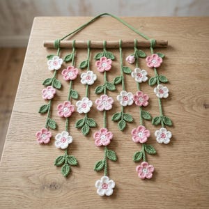 Op de afbeelding: Een handgemaakte wandhanger met gehaakte roze en witte bloemen en groene bladeren. De bloemen zijn opgehangen aan een houten deuvel met groen garen. Dit decoratieve stuk voegt een vleugje door de natuur geïnspireerde charme toe.
