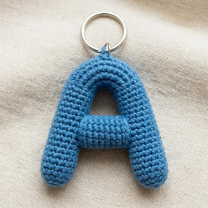 Puede incluir: Un llavero con la letra A tejida a ganchillo en color azul, con una superficie texturizada de hilo y una anilla de metal plateado en la parte superior. La letra está construida con un patrón de puntos denso y uniforme, creando una forma tridimensional.