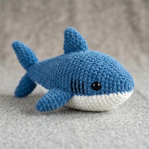 Puede incluir: Un peluche de tiburón tejido a mano en azul y blanco. El tiburón tiene un aspecto texturizado y suave con detalles negros en los ojos y las branquias. El peluche está sobre una superficie gris.