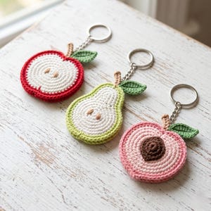 May include: Three crochet fruit keychains: a red apple, a green pear, and a pink peach, each with a white interior, brown seeds, and green leaves. The keychains are attached to silver key rings.
