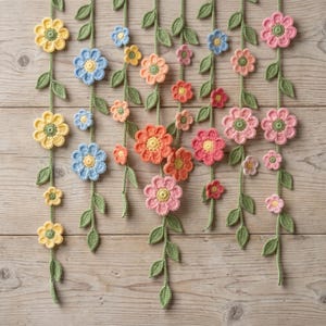 Op de afbeelding: Een verzameling gehaakte bloemen in verschillende kleuren, waaronder geel, blauw, oranje en roze, gerangschikt op groene ranken met bladeren. De bloemen zijn van verschillende grootte en staan tegen een lichtbruine houten achtergrond.