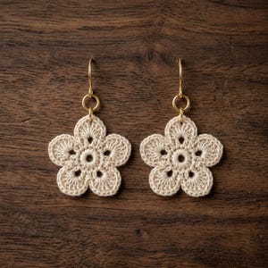 May include: A pair of cream-colored, crocheted flower earrings with gold-colored ear wires. The earrings are handmade and feature a five-petal design with a circular center. The background is a dark brown wooden surface.