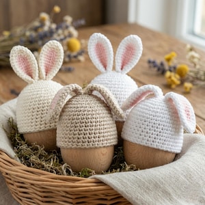 May include: A wicker basket holds four decorative Easter eggs. Each egg has a wooden base and is topped with a crocheted bunny hat. The hats are in cream, tan, and white, with pink inner ears. The basket sits on a wooden surface.