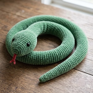 May include: A green crocheted snake plush toy with a textured yarn surface, featuring two small black plastic eyes and a bifurcated red yarn tongue protruding from its mouth. The snake is coiled into a circular shape on a wooden surface.
