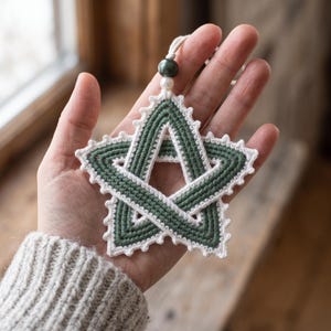 Könnte beinhalten: Ein fünfzackiger Sternschmuck aus dunkelgrünem und weißem Häkelgarn. Der Stern hat ein verschlungenes Design mit einem weißen Rand aus Picots. Eine dunkelgrüne Perle und eine kleine weiße Perle sind an der oberen Schlaufe befestigt.