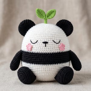 Pode incluir: Um peluche de panda em croché com formato redondo, corpo branco, secção central preta, orelhas pretas e membros pretos. O panda tem olhos fechados, um pequeno nariz preto e bochechas cor-de-rosa em forma de flor. Um pequeno rebento verde está no topo da cabeça.