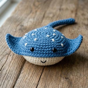 Könnte beinhalten: Handgefertigter blauer Häkelrochen mit weißen Punkten und schwarzen Augen. Der Rochen hat einen cremefarbenen Bauch und ein lächelndes Gesicht. Das Spielzeug liegt auf einer rustikalen Holzoberfläche.
