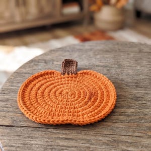 May include: A round, orange crocheted coaster shaped like a pumpkin, featuring a small, rectangular brown crocheted stem at the top center. The coaster is constructed with concentric circular stitches in a vibrant orange yarn, creating a textured surface.