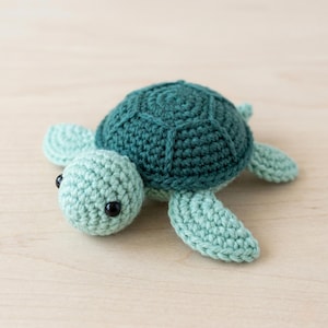 May include: A crocheted turtle toy in teal and seafoam green. The turtle features a textured shell, a rounded head, and four flipper-like limbs. Black safety eyes give the turtle a friendly appearance. A charming, handcrafted item.