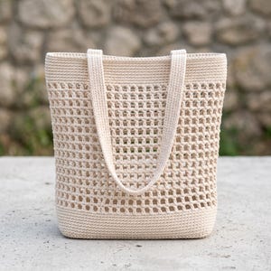 Op de afbeelding: Een crèmekleurige tote bag met een dichte, stevige bodem en een wijd, open gehaakt patroon over het hoofdgedeelte. De tas is voorzien van twee bijpassende crèmekleurige schouderbanden die stevig aan de bovenrand zijn bevestigd.