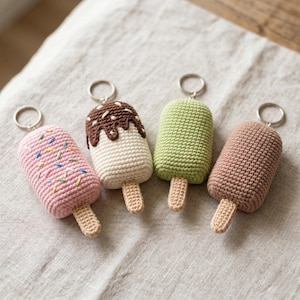 Puede incluir: Cuatro llaveros de paletas de helado hechos a mano con ganchillo en rosa, blanco, verde y marrón. La paleta rosa tiene chispas de colores. Cada llavero tiene un palito de madera y un anillo de metal plateado.