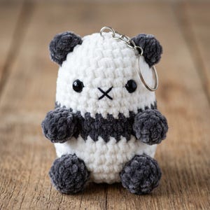 May include: A handmade crochet panda keychain. The panda has a white body with black accents, including the ears, paws, and a stripe around the middle. It features black button eyes and a stitched X for a mouth. A silver keychain ring is attached.