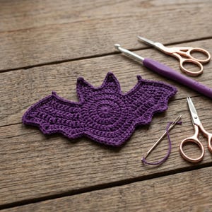 May include: A handcrafted, purple crochet bat sits on a wooden surface, accompanied by a crochet hook, scissors, and a needle with thread. The bat features a circular body and pointed wings, ideal for Halloween crafts.