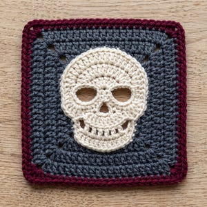 May include: A square crochet coaster featuring a central off-white skull motif on a dark gray background, framed by a deep burgundy border. The piece is crafted with visible yarn stitches, creating a textured surface with a clear skull design.