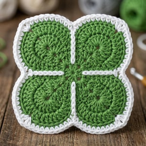 Puede incluir: Un posavasos de ganchillo hecho a mano con forma de trébol de cuatro hojas. El posavasos presenta un centro verde vibrante con un borde blanco. El diseño del trébol está detallado con puntadas visibles, mostrando la artesanía.