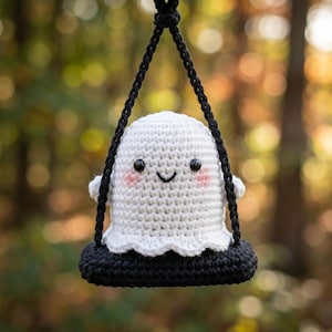May include: A crocheted white ghost with a smiling face and rosy cheeks, hanging from a black swing. The ghost has small arms and a scalloped bottom edge. The swing is suspended by black cords against a blurred background of green and gold.