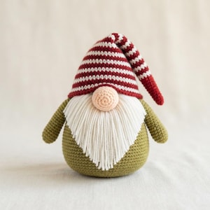 May include: A crochet gnome figurine featuring an olive green rounded body and arms. It wears a tall, pointed hat with horizontal red and white stripes that flops to one side. A long, white yarn beard covers the front, with a small peach-colored nose.