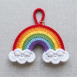 Può includere: Arcobaleno all'uncinetto fatto a mano, con arco superiore rosso, bande arancioni, gialle, verdi, blu e viola. Nuvole bianche in basso. Anello rosso con anello argentato in alto. Artigianato decorativo.